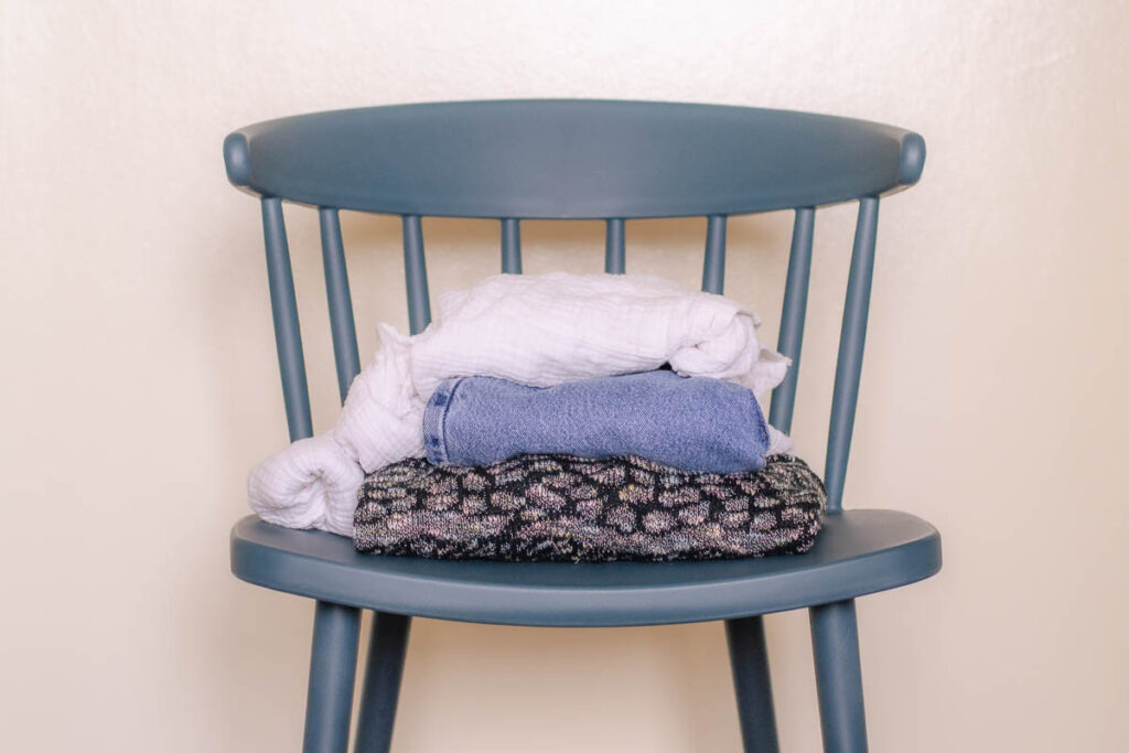 Blue chair that holds a white blouse, blue jeans, and sweater that are folded and piled on top of each other